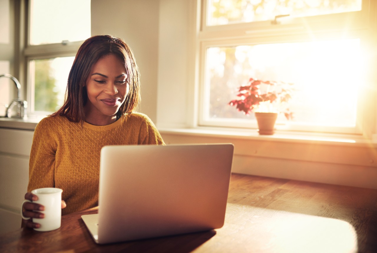 Adult female holding cup and laughing while looking at her laptop computer while sitting near bright sunny window in kitchen