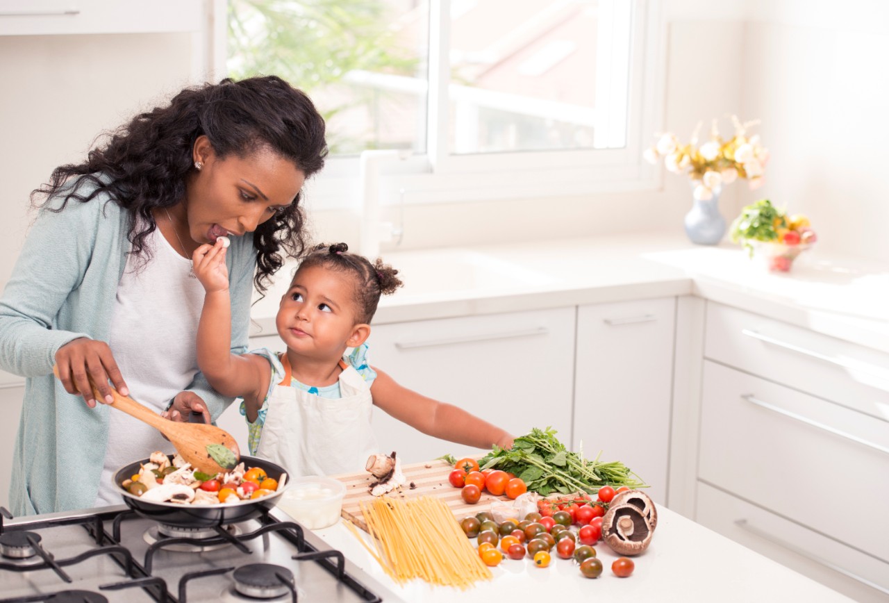 Mother with her 2.5 years little girl daughter preparing pasta with red sauce together. Woman and girl using fresh tomato, mushrooms, basil and garlic.