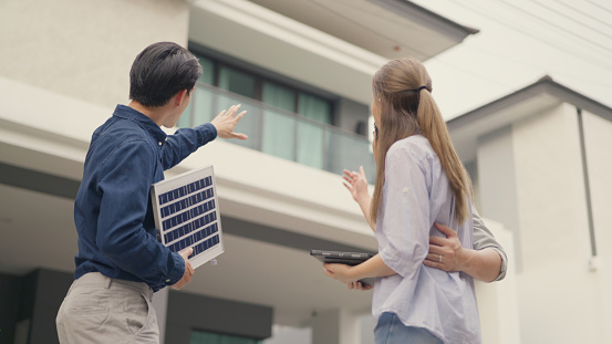 A solar energy expert holding a panel discusses efficient power solutions with a couple in front of their residential building, outlining the benefits of renewable energy.